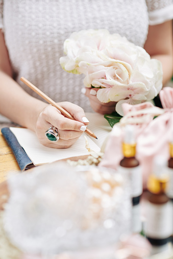 Hands of woman with light pink flower in hand writing ideas for next organic cosmetics line under her brand