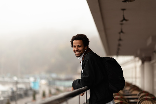 Positive african man with backpack smiling, foggy weather.