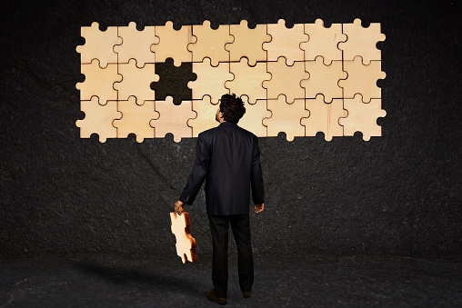 A businessman holding the missing piece of puzzle to an unfinished puzzle hanging on a black textured wall