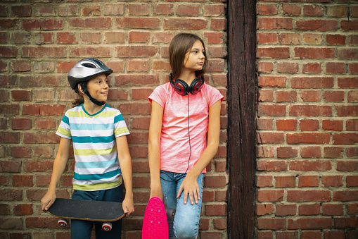 Boy and girl leaning on brick wall and holding their skateboards. They are beautiful and happy. Boy has a protective sport helmet on his head