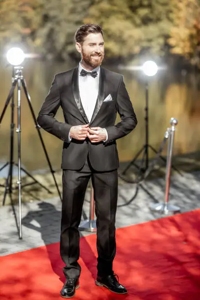 Full body portrait of an elegant man strictly dressed in tuxedo posing on the red carpet outdoors