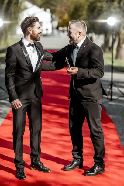 Two elegant men strictly dressed in suits as a well-known film actors having fun during awards ceremony on the red carpet outdoors