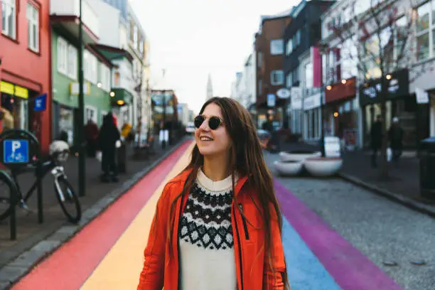 Young woman with long hair, in sunglasses, Icelandic style wool sweater and red jacket walking on the colorful streets of Reykjavik in Iceland, enjoying the winter vibes and Christmas decoration, smiling and playing with a small ball