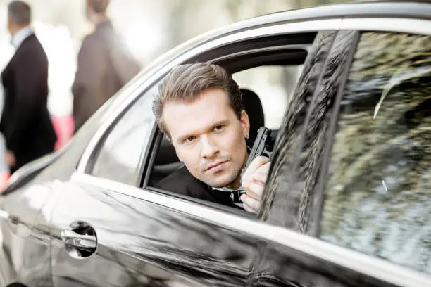 Portrait of a man as a famous movie actor sitting with handgun in the luxury car, arriving on the awards ceremony near the red carpet outdoors
