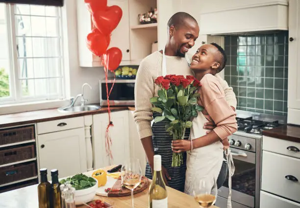 Cropped shot of an affectionate young couple posing with a bunch of flowers in their kitchen on their anniversary