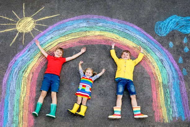 Photo of three little children, two school kids boys and toddler girl having fun with with rainbow picture drawing with colorful chalks on asphalt. Siblings in rubber boots painting on ground playing together.