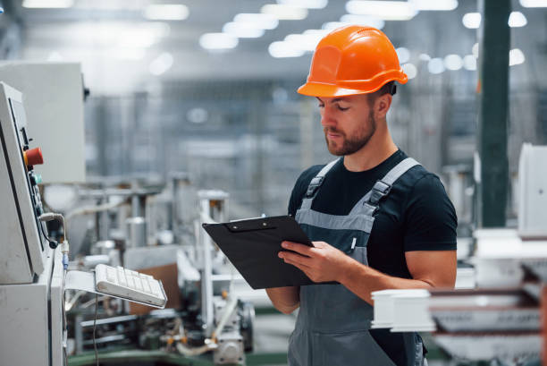 Looks at equipment and makes notes. Industrial worker indoors in factory. Young technician with orange hard hat Looks at equipment and makes notes. Industrial worker indoors in factory. Young technician with orange hard hat. industrial orange stock pictures, royalty-free photos & images