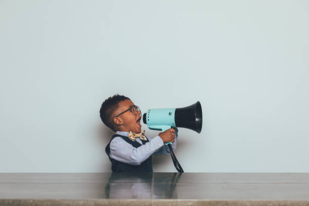 Young Nerd Boy with Megaphone A young nerd boy dressed in bow tie, vest and wearing eyeglasses, holds and yells through a megaphone trying to get someone to listen to him. He sits at a table trying to get his message across to others. black nerd stock pictures, royalty-free photos & images