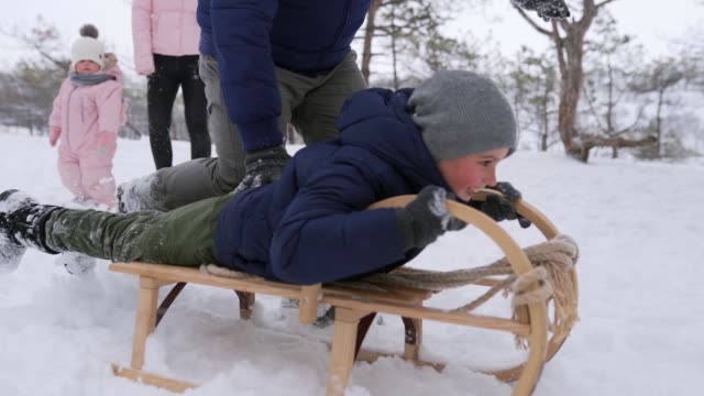 Father launches son pushing sled down the hill on snowy winter day in park. Boy sledge outdoors. Happy family sledding. People sleigh ride and enjoy Christmas vacation in forest. Slow motion