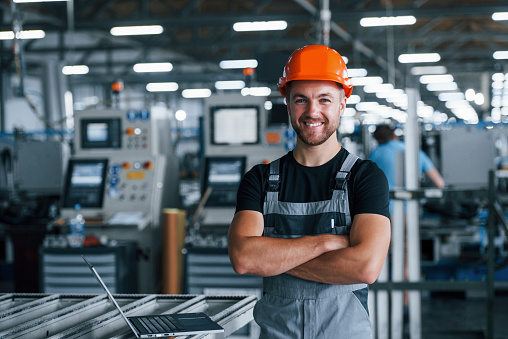 Smiling and happy employee. Industrial worker indoors in factory. Young technician with orange hard hat.