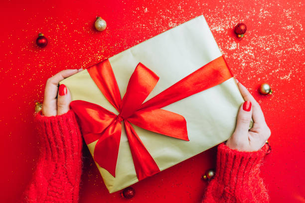 istock Female hands holding present with red bow on red sparkling background. Festive backdrop for Christmas or New Year. Flat lay. Top view 1186946493