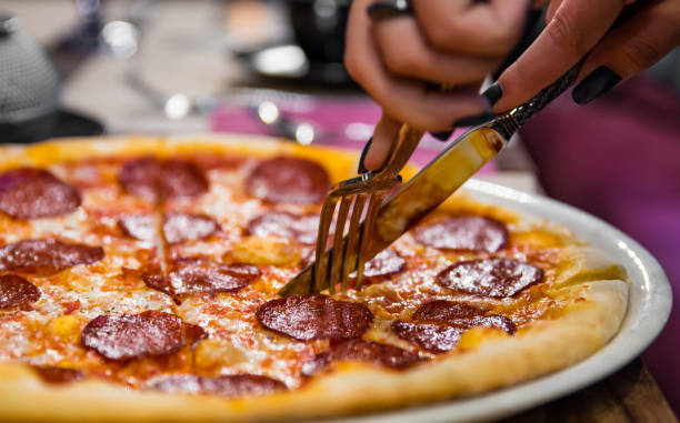 woman hands with knife and fork cutting pepperoni pizza on table in cafe