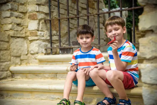 Photo of The children playing on the ruins of ancient building with metal gate an archaeological site of an ancient city. Two boys sitting and play with toy aircraft plane. Travel concept