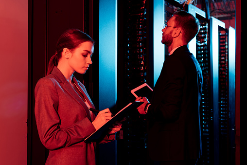 attractive businesswoman holding clipboard near businessman with digital tablet in data center
