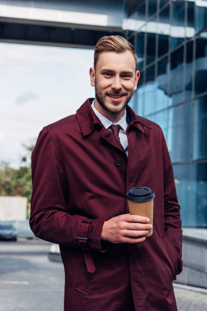istock happy bearded man in coat holding paper cup while standing with hand in pocket 1186956256