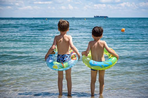 Two cute little brother boy stand on the beach in swimming trunks goes swimming with inflatable ring. Safe water activities with kids.