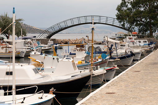 Chios, Greece - July 14, 2019: The harbor of the Lagada village situated on the east coast of the Greek Island of Chios in the Aegean Sea. Line of boats are tied up (or moored) at the quarry (or pier), including fishing boats, sailing boats and recreational motor boats. A bridge crosses the water at the entrance of the harbor or marina.