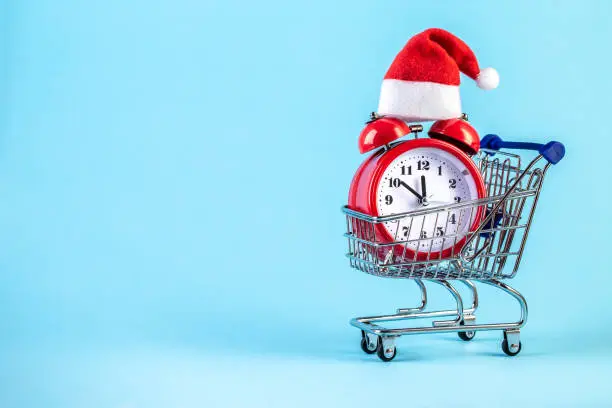 Christmas alarm clock in a supermarket trolley on a blue background. Alarm clock with Santa Claus hat. Merry Christmas and Happy New Year concept. Template mockup for greeting card your text design