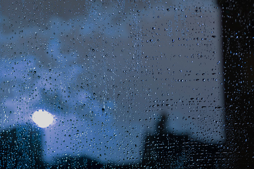 Wet window glass with lots of rain drops and a view of the night gray sky with the white glowing moon and blurred dark silhouettes of houses in the pale moonlight