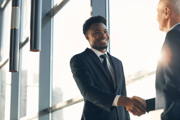istock Cheerful handsome young Black businessman in suit standing at panoramic window and making handshake with business partner in lobby of company 1186986873
