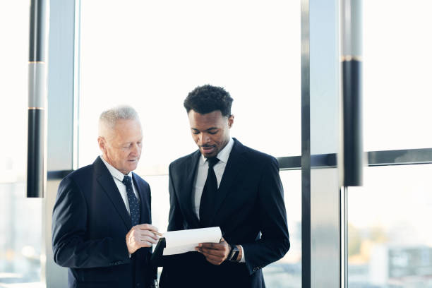 istock Two multi-ethnic businessmen in formal suits standing against panoramic window and analyzing papers together in office 1186986883