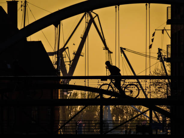 Silhouette of a biker an a Hamburg bridge at sunset. Industrial setting with cranes and port structures in the background.