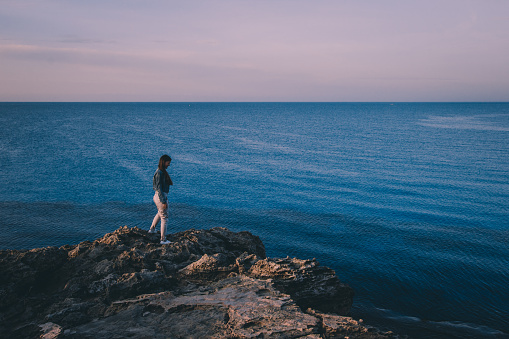 Woman traveler standing near sea on top of cliff in summer mountains and enjoying view of nature and sea. Cape Greco, Cyprus, Mediterranean Sea. Sunrise.