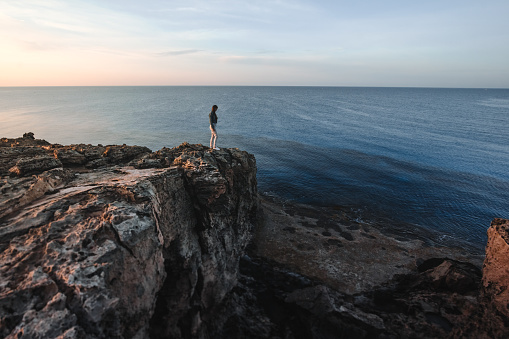 Woman traveler standing near sea on top of cliff in summer mountains and enjoying view of nature and sea. Cape Greco, Cyprus, Mediterranean Sea. Sunrise.