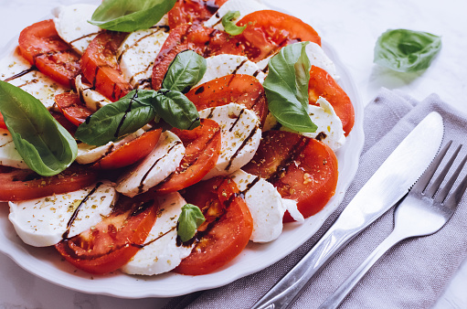 Plate of healthy classic delicious caprese salad with ripe tomatoes and mozzarella cheese with fresh basil leaves on white marble background. Italian cuisine concept.