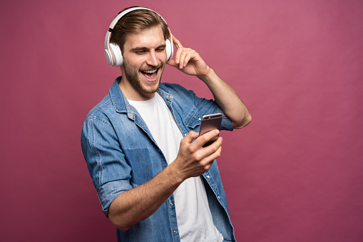 Excited young man wearing jeans shirt standing isolated over pink background, listening to music with earphones and mobile phone