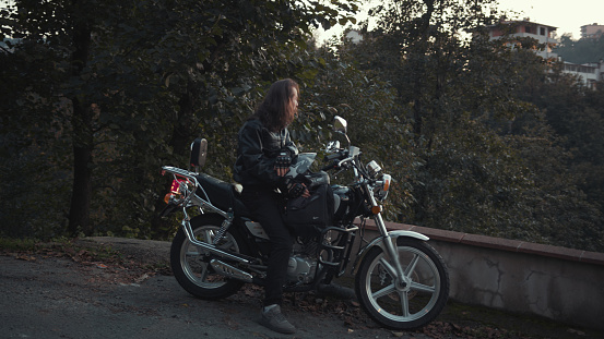 Handsome biker man with long hair wearing a leather jacket sitting on his motorcycle looking at the sunset. Vacation concept.