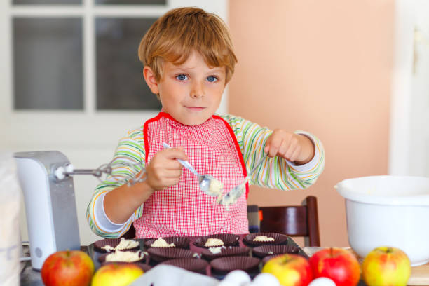 cute little happy blond preschool kid boy baking apple cake and muffins in domestic kitchen. funny lovely healthy child having fun with working with mixer, flour, eggs, fruits. little helper indoors - cake making mixing eggs imagens e fotografias de stock