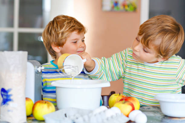 two little funny brothers baking apple cake in domestic kitchen. happy healthy kid boys having fun with working with mixer, eggs and fruits. children tasting dough indoors - cake making mixing eggs imagens e fotografias de stock