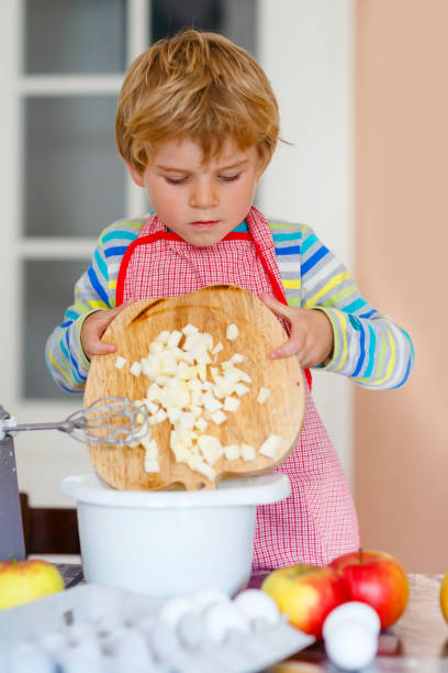 cute little happy blond preschool kid boy baking apple cake and muffins in domestic kitchen. funny lovely healthy child having fun with working with mixer, flour, eggs, fruits. little helper indoors - cake making mixing eggs imagens e fotografias de stock