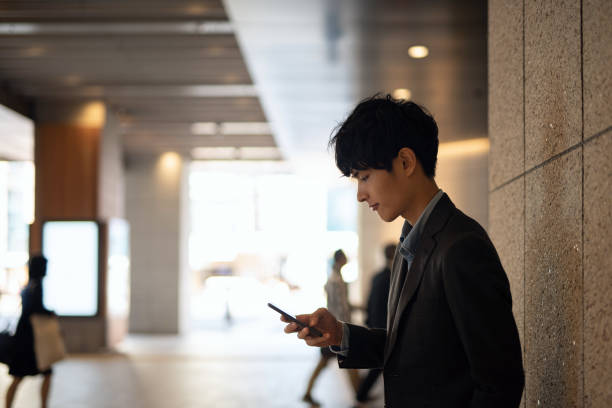 歩行者歩道でスマートフォンを見ている若いビジネスマン - スマホ 日本人 ストックフォトと画像