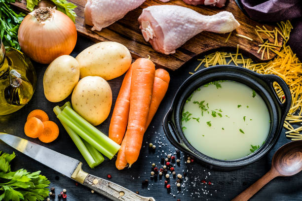Ingredients for cooking chicken broth Top view of a cooking pan filled with broth surrounded by the ingredients for cooking a chicken soup like chicken thigh, noodles, olive oil, celery, carrots, potatoes, onion and parsley. Low key DSLR photo taken with Canon EOS 6D Mark II and Canon EF 24-105 mm f/4L chicken soup stock pictures, royalty-free photos & images