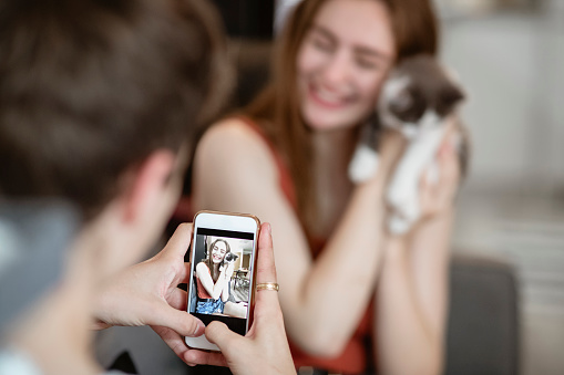 Male taking a picture of his girlfriend holding their pet kitten on his phone.
