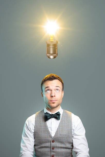 Man with a great idea A man with a bow tie and waistcoat is looking towards a glowing light bulb directly above his head. man with a light bulb over his head stock pictures, royalty-free photos & images