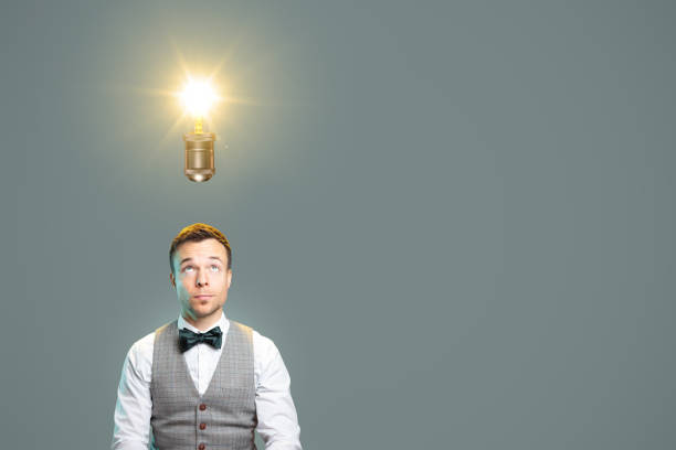 Man having a big idea A man with a bow tie and waistcoat is looking towards a glowing light bulb directly above his head. Copy space to the right. man with a light bulb over his head stock pictures, royalty-free photos & images