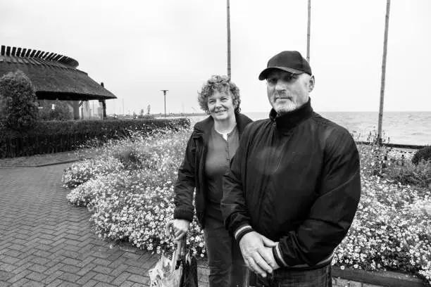 Nida, Lithuania - Sept 8, 2019: happy elderly married couple standing and looking at camera with warm clothes on and holding umbrellas on the coastline path at he Baltic sea.