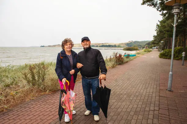 Nida, Lithuania - Sept 8, 2019: happy elderly married couple standing and looking at camera with warm clothes on and holding umbrellas on the coastline path at he Baltic sea.