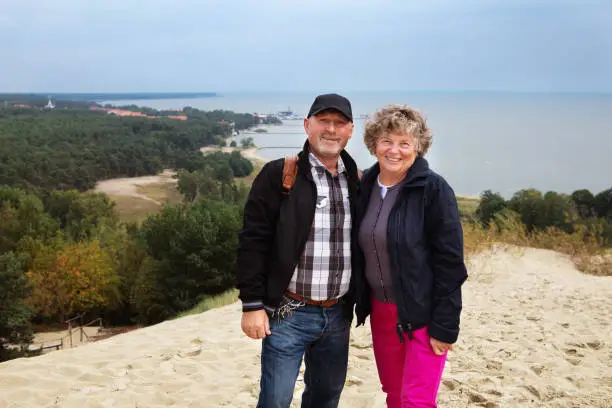 Nida, Lithuania - Sept 8, 2019: elderly couple smiling standing and looking at camera while posing with a beautiful scenery of the Baltic sea and Nida village behind them.