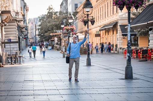 Mature Confident Businessman with Grey Hair and Beard is Walking in the City Center and Greets Someone with Arm Raised. Older Successful Man in Blue Shirt and Sunglasses is Walking Down the City Streets with his Briefcase.