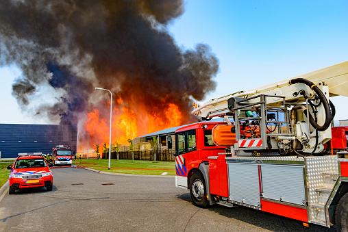 Fire engines in front of a large fire in a factory in an industrial area. Huge flames and thick smoke are emerging from the warehouse.
