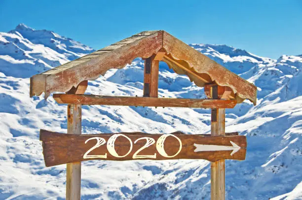 Photo of 2020 written on a wooden direction sign, snow mountain landscape on the background, ski resort holiday greeting card