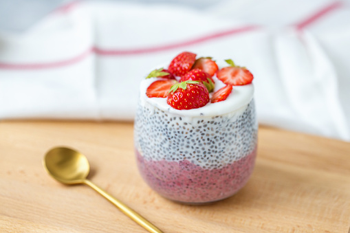 Chia seed pudding with almond milk, yogurt, pink pitaya dragon fruit powder and strawberries dessert in a glass, spoon. Superfood and vegan food concept. Copy space, selective focus.