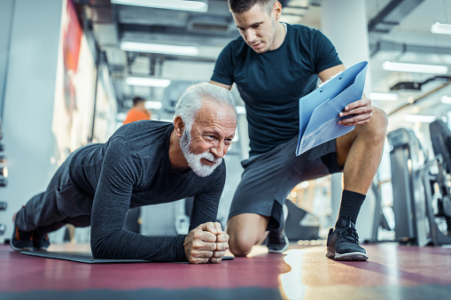 Active mature man exercising strength in plank position while his young coach is next to him. Copy space.