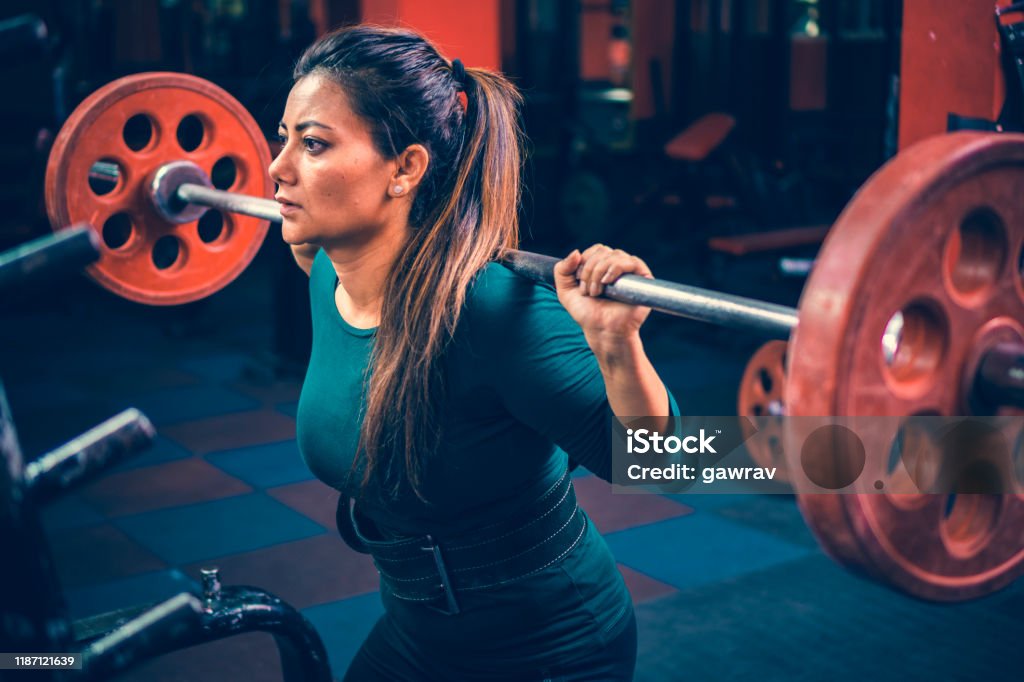 Woman does squats workout with a barbell in a gymnasium for fitness. Indoor image of an Asian/Indian, mid-adult woman does squats workout with a barbell in a gymnasium for her fitness. Indian Ethnicity Stock Photo
