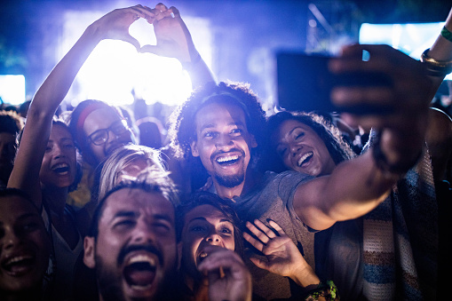 Large group of happy friends having fun while taking a selfie with cell phone on music concert.