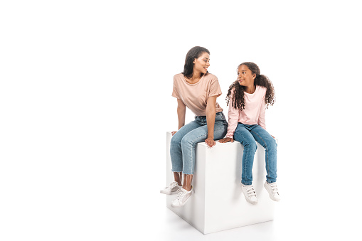 smiling african american mother and daughter looking at each other while sitting on white cube on white background